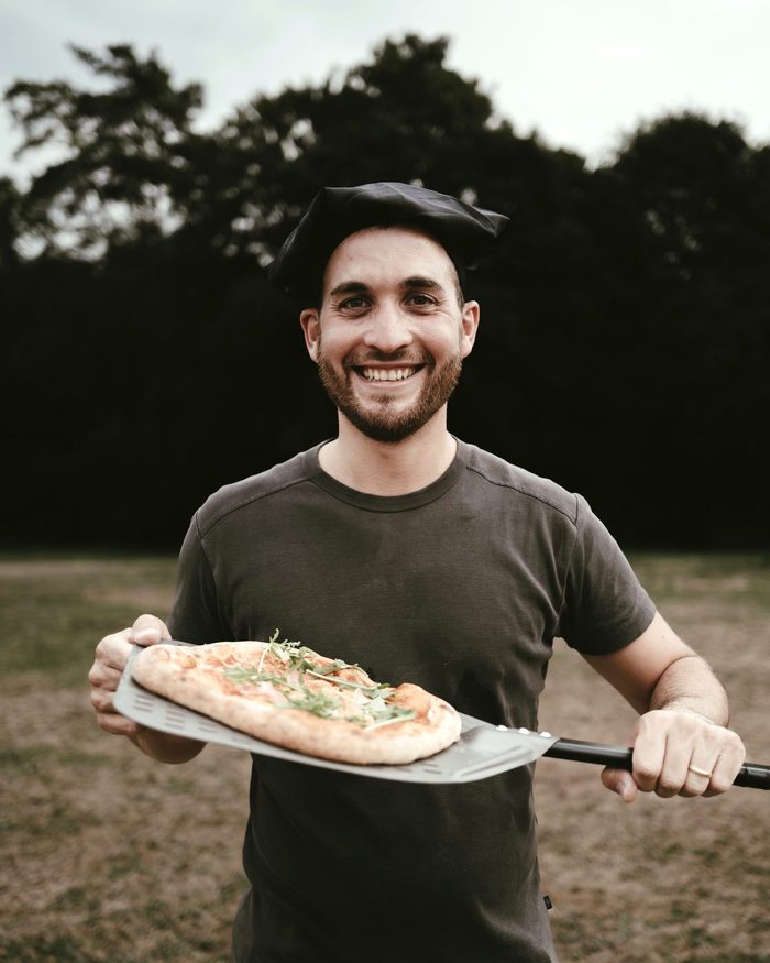 Silas Gärtig mit Kochmütze hält eine frisch gebackene Pizza auf dem Pizzaschieber